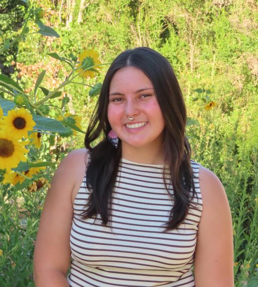 Smiling young woman with long dark hair in a striped top by sunflowers and green foliage.